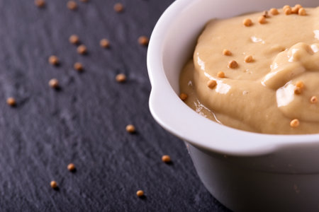 Horizontal photo of white bowl full of mustard. Fresh orange mustard in bowl with several seeds on surface. Mustard seeds spilled around white bowl. Bowl with mustard on black slate stone.の写真素材
