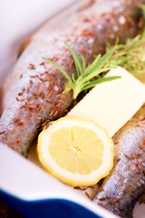 Vertical photo of two raw trouts. Fishes in blue ceramic pan on wooden board with green towel around. Herbs as thyme and rosemary with butter and lemon slices together with meat.の写真素材