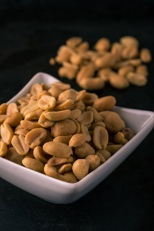Vertical photo of salted peanuts. White square full of nuts with salt in front of heap of others. Nuts placed on dark black baking tray board with worn surface.の写真素材