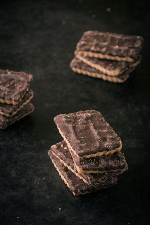 Vertical photo with few stacks of square biscuits with chocolate on one side. Each stack consists of four or free pieces. Sweets are placed on dark black baking tray with textrured worn surface.の写真素材