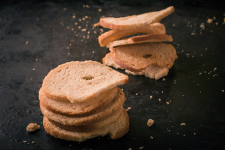 Horizontal photo of two stacks created by baked rolls cut to slices with different shapes. Several crumbs are spilled around on dark black baking tray with worn rustic surface.の写真素材