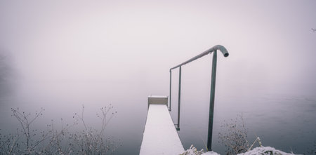 Horizontal photo of winter landscape with heavy morning fog. Footbridge with metal railings goes partially over the pond covered by ice. Fog coveres a background. Grass is in front of pic.の写真素材