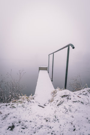 Vertical photo of winter landscape with heavy morning fog. Footbridge with metal railings goes partially over the pond covered by ice. Fog covers a background. Grass is in front of pic.の写真素材