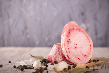 Horizontal photo with two pieces or slices of big marrowbone with few drops of blood and meat on side. Bones are placed on crumpled paper several spices. All is on wooden board with white color.の写真素材