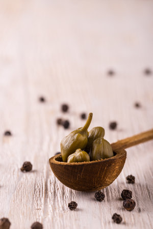 Vertical photo of pickled green capers. Few pieces of edible blooms are placed in wooden vintage spoon. Several seeds of whole pepper spice are spilled around on light white wooden board.の写真素材
