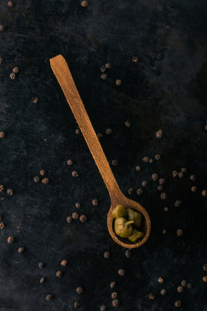 Vertical photo with top view on pickled green capers. Few pieces of edible blooms are placed in wooden vintage spoon. Several seeds of whole pepper spice are around on dark worn baking tray.の写真素材