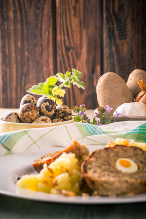 Vertical photo of yellow bowl full of quail eggs next to portion of Easter meatloaf filled by eggs with meshed potatoes and pan with vegetable. Few flowers and herbs are around the food.の写真素材