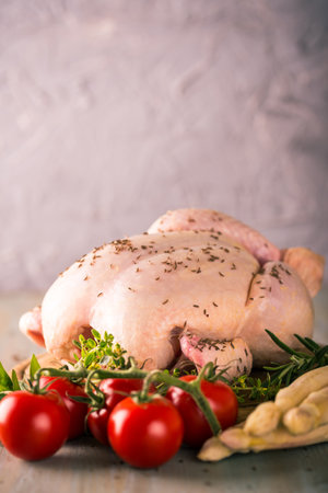 Vertical photo of whole raw chicken. Meat has a spice on a skin like pepper or cumin. Few herbs are around as thyme and rosemary. Chicken is on rounded wooden board with vegetable.の写真素材
