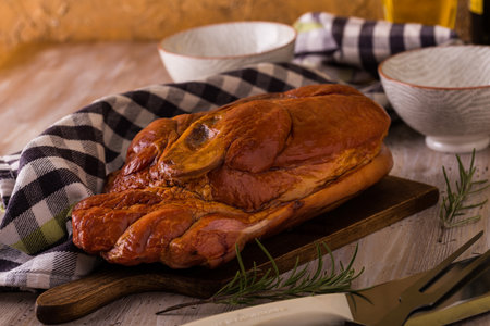 Horizontal photo with big piece of smoked pork meat with bone placed on cutting board. Blue towel, chopper, fork, bowls and rosemary are around with wine and oil in background with vintage wall.の写真素材