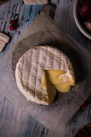Vertical photo with top view on single roasted camembert with white skin. Small piece is cut off and the cheese is partially melted. Cheese is on paper sheet and blue vintage wooden board.の写真素材