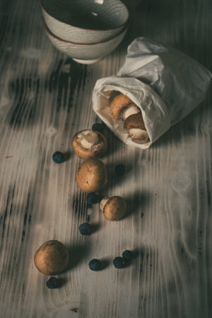 Vertical vintage photo of several brown edible mushrooms. Champignons are spilled on white wooden board from paper bag. Few dark blue backthorn berries are around with bowls and bottles.の写真素材