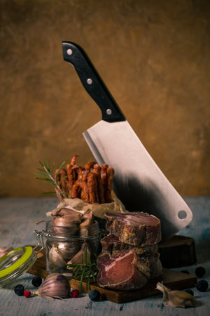 Vertical photo with raw dried and smoked tuscany meat from wild boar. The meat is cut to big pieces and stacked on vintage wooden board with garlic in glass and sausages in can.の写真素材