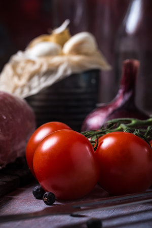 Vertical photo of few red cherry tomatoes. The vegetable is placed on vintage wooden board next to raw pork tenderloin meat, purple onion and old can full of white garlic cloves.の写真素材
