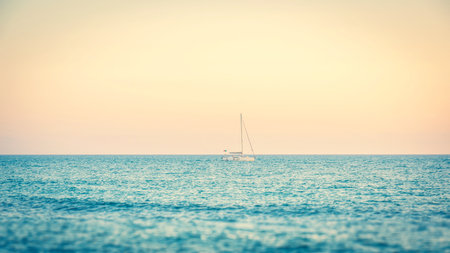 Horizontal photo with small ship. Boat has white color and high mast. Boat is alone on surface of blue sea. Scene is captured during dusk. Sky is orange with few clouds.の写真素材