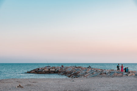 Horizontal photo with stone breakwater. Breakwater goes to blue sea. Sand beach is in front of sea. Clear evening sky is with few clouds. Several persons are on the structure.の写真素材