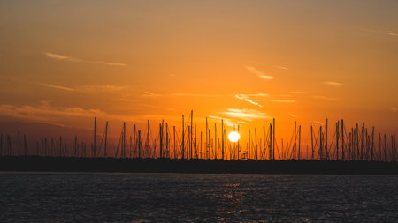 Horizontal photo of dusk. Evening sky has orange color and few clouds. Sun is near horizon. Many ships with silhouettes of masts are in harbour. Dark see with small curls is in front.の写真素材