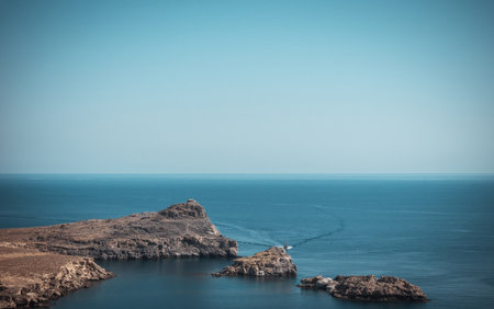 Horizontal photo with dry rocks covered just by stones. Rocks are brown and grey. Rocks are next to famous town Lindos on Rhodes island. Sky and sea are blue.の写真素材