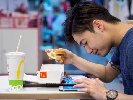 SINGAPORE - 18 MAR 2019 - A young man eats a McDonalds meal while using his smartphoneのeditorial素材