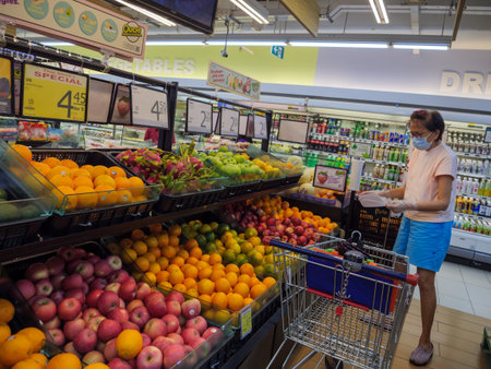 SINGAPORE â 2 APR 2020 â Middle aged woman wearing surgical mask shops for groceries at an NTUC Fairprice supermarket. The supermarket shelves are full of fruits â apples, oranges, dragonfruit, canned drinks and beveragesのeditorial素材