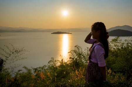 Happy young woman standing on the hill watching the sunset over the lake in Kaeng Kra Chan National Park, Phetchaburi Province, Thailandの写真素材