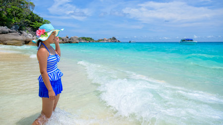 Beautiful woman in a blue striped swimsuit and hat standing poses on beach of sea under a summer sky at Koh Miang Island in Mu Ko Similan National Park, Phang Nga Province, Thailand, 16:9 widescreenの写真素材