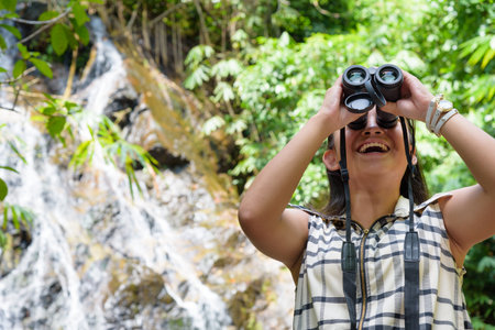 Beautiful young girl hiking is using binoculars look for birds in tropical forest near the waterfalls in Thailandの写真素材