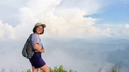 Teens girl hiker wear a caps and glasses with backpack is standing and smiling happily on high mountain at scenic point of Phu Chi Fa Forest Park in Chiang Rai Province Thailand, 16:9 widescreenの写真素材