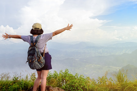 Happy teens girl hiker with backpack and caps open arms looking beautiful nature landscape on high mountain at viewpoint of Phu Chi Fa Forest Park during sunset in Chiang Rai Province Thailandの写真素材