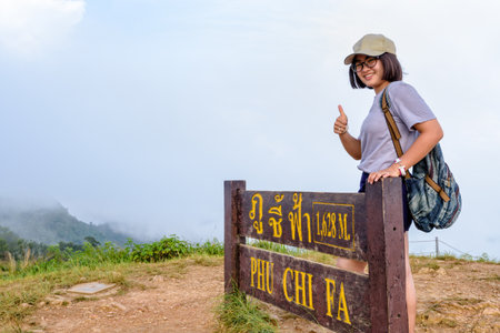 Tourist teen girl hiker wear caps and glasses with backpack smiling thumb up near nameplate attractions of Phu Chi Fa Forest Park on mountain and mist background, Chiang Rai, Thailandの写真素材