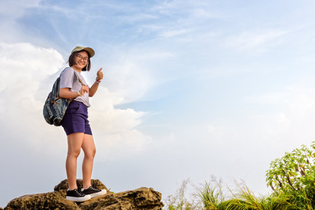 Happy hiker asian cute teens girl with backpack caps and glasses standing smiling poses thumb up on mountain and sky background at Phu Chi Fa Forest Park during sunset in Chiang Rai, Thailandの写真素材