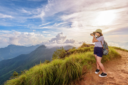 Hiker asian teen girl wear cap and eyeglass with happy holding dslr camera shooting photo beautiful nature in winter on peak mountain at sunset in Phu Chi Fa Forest Park, Chiang Rai, Thailandの写真素材