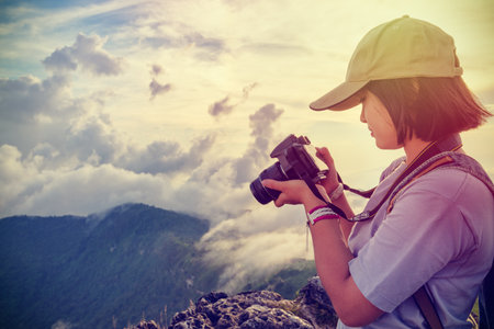 Vintage style hiker teens girl wear caps looking photo at digital camera is beautiful landscape natural of sierra and sky during sunset on mountain in Phu Chi Fa Forest Park, Chiang Rai, Thailandの写真素材
