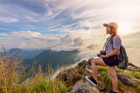 Hiker asian young woman happy with camera backpack and caps looking beautiful landscape nature of mountain and colourful sky at sunset on viewpoint Phu Chi Fa Forest Park, Chiang Rai, Thailandの写真素材