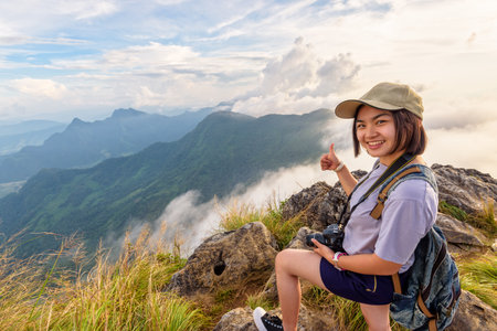 Happy hiker asian cute teens girl with dslr camera caps and backpack stand smiling poses thumb up on top of mountain at viewpoint Phu Chi Fa Forest Park in winter, Chiang Rai, Thailandの写真素材