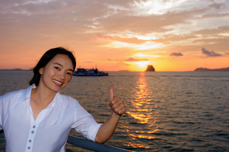 Women raise their thumb to admire the beautiful nature of colorful sky and sun at sunset over the sea on the deck of a passenger boat while cruising to Koh Samui Island in Surat Thani, Thailandの写真素材