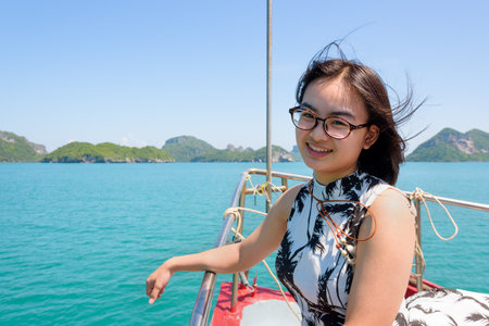 Cute young woman with eyeglasses smiling happily on the prow boat while cruising the beautiful natural of the blue sea and sky in summer at Mu Ko Ang Thong National Park, Surat Thani, Thailandの写真素材