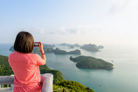 Woman tourist on peak viewpoint of Ko Wua Ta Lap island use camera take photos at beautiful nature landscape during sunrise over the sea in Mu Ko Ang Thong National Marine Park, Surat Thani, Thailandの写真素材