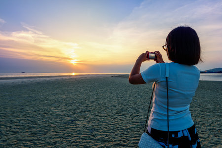Asian woman tourist taking photo on the beach and beautiful natural landscape of colorful sky and sea during a sunset at Nathon Sunset Viewpoint in Samui island, Surat Thani, Thailandの写真素材