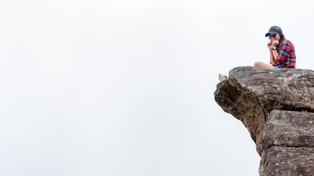 Asian woman sitting on a rock near a cliff on a white fog background is pretending to be thrilled, 16:9 widescreenの写真素材