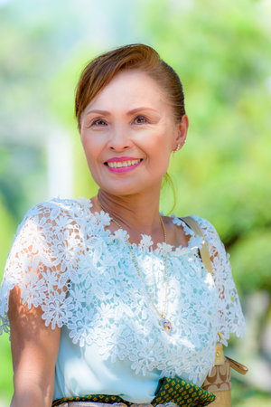 Front portrait of beautiful Asian middle-aged woman in good health, Dress in Thai style clothes smiling happily on the nature green tree backgroundの写真素材
