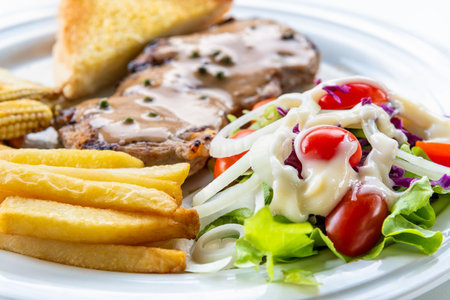 Closeup side view vegetable salad topped mayonnaise with tomato, onion, lettuce, purple cabbage and steak pork chop with pepper sauce and french fries on a clean white dish for cooking delicious mealsの写真素材