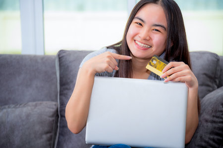 Asian young woman holding SME credit card use a laptop computer buy products payment shopping online, Cute teenage girl smile pointing finger at credit card happy lifestyle through credit card paymentの写真素材