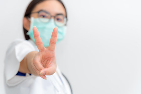 Studio portrait Asian young woman doctor closeup raise two V shape finger wear a mask to safety Coronavirus to support in the fight against disease epidemic COVID 19 concept on blank gray backgroundの写真素材