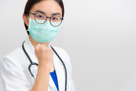Beautiful Asian young woman doctor in white uniform and mask protect Coronavirus, Physician raise fist to support patient in the fight against disease epidemic COVID 19, gray copy space backgroundの写真素材