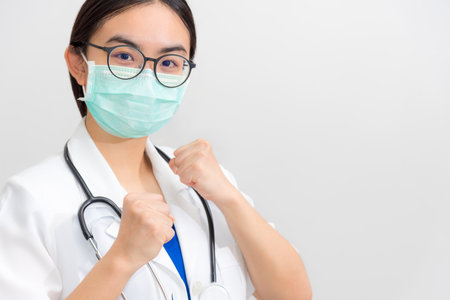 Beautiful Asian young woman doctor in white uniform and mask protect Coronavirus, Physician raise fists to support patient in the fight against disease epidemic COVID 19, gray copy space backgroundの写真素材