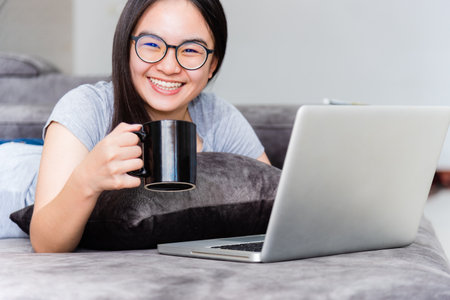 Portrait beautiful Asian young woman holding coffee cup smile and looking during a work, Cute girl teens happy on the sofa working remotely with a laptop and internet computer communication from homeの写真素材