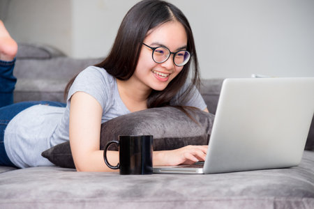 Portrait beautiful Asian young woman lying relax work with a laptop, Cute girl teens looking at the monitor smile happily on the sofa working remotely with an internet computer communication from homeの写真素材