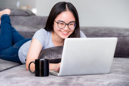 Portrait beautiful Asian young woman lying relax work with a laptop, Cute girl teens looking at the monitor smile happily on the sofa working remotely with an internet computer communication from homeの写真素材