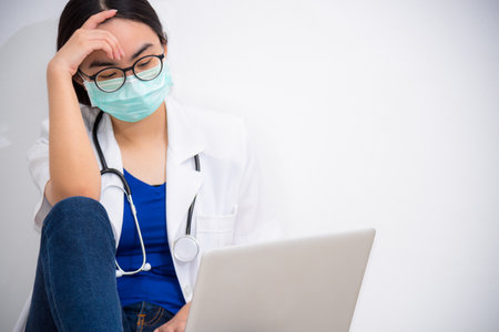 Asian young woman doctor wear mask to safety Coronavirus sit on the floor looking at the laptop with sadness, stress and worry about the problem, Concept take care yourself stop the epidemic Covid-19の写真素材