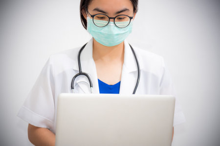 Studio portrait Asian young woman doctor wears glasses and mask to safety Coronavirus looking at the laptop, Physician with stethoscope are working on a computer to stop the epidemic Covid-19 virusの写真素材